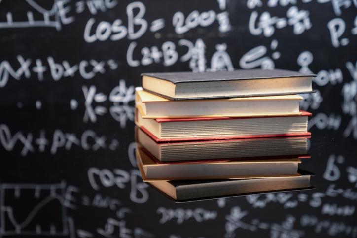 Textbooks stacked on wooden school desk in front of green chalkboard. Classroom setting.